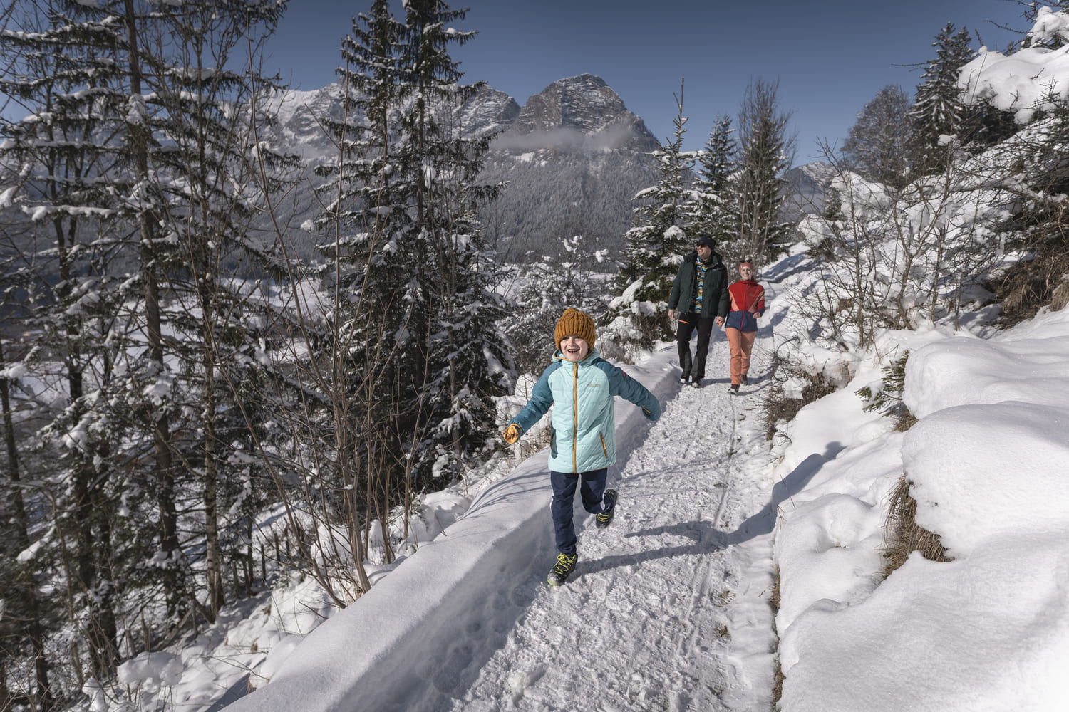 Kind mit orangefarbener Strickmütze und hellblauer Jacke läuft lachend auf verschneitem Bergweg, zwei Begleitpersonen folgen.