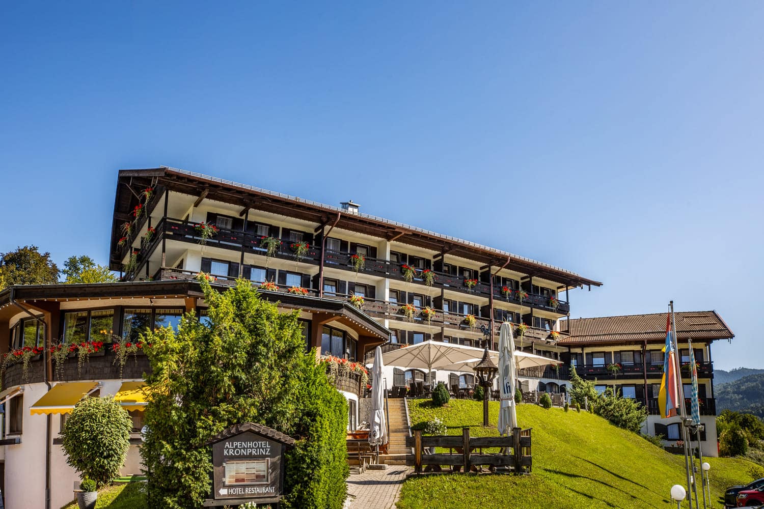 Alpenhotel Kronprinz, großes Alpenhotel auf sonnigem Hügel mit Balkonen, Blumenampeln und Terrasse mit Schirmen.