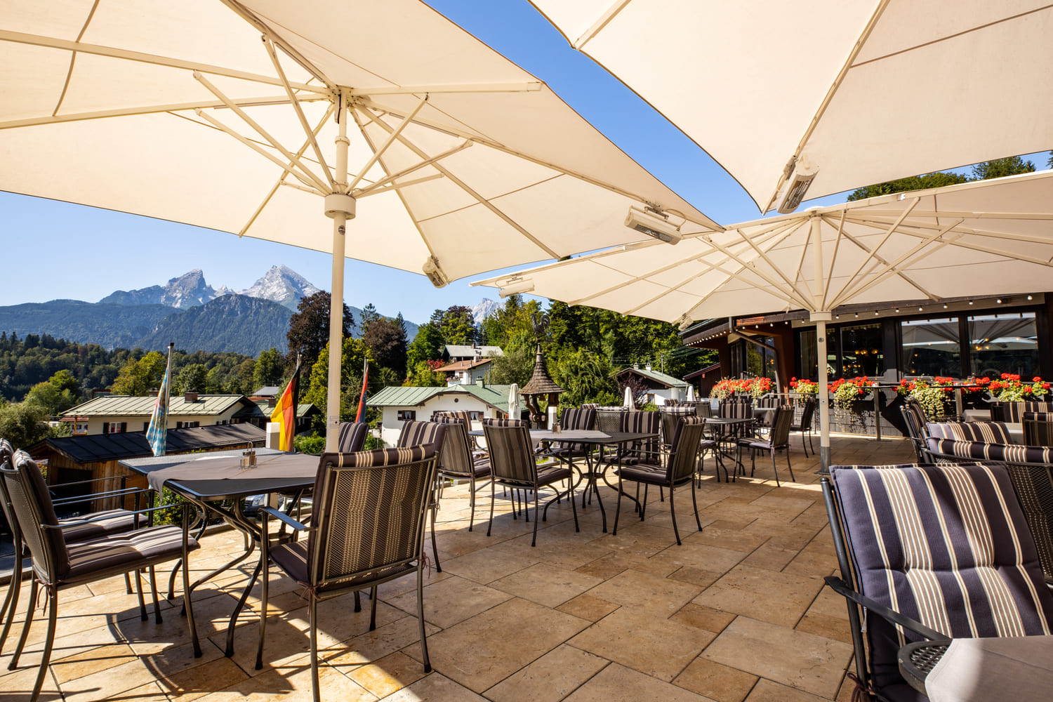 Sonnige Terrasse mit gestreiften Sitzpolstern und großen weißen Sonnenschirmen, Blick auf bewaldete Hügel und Berge.