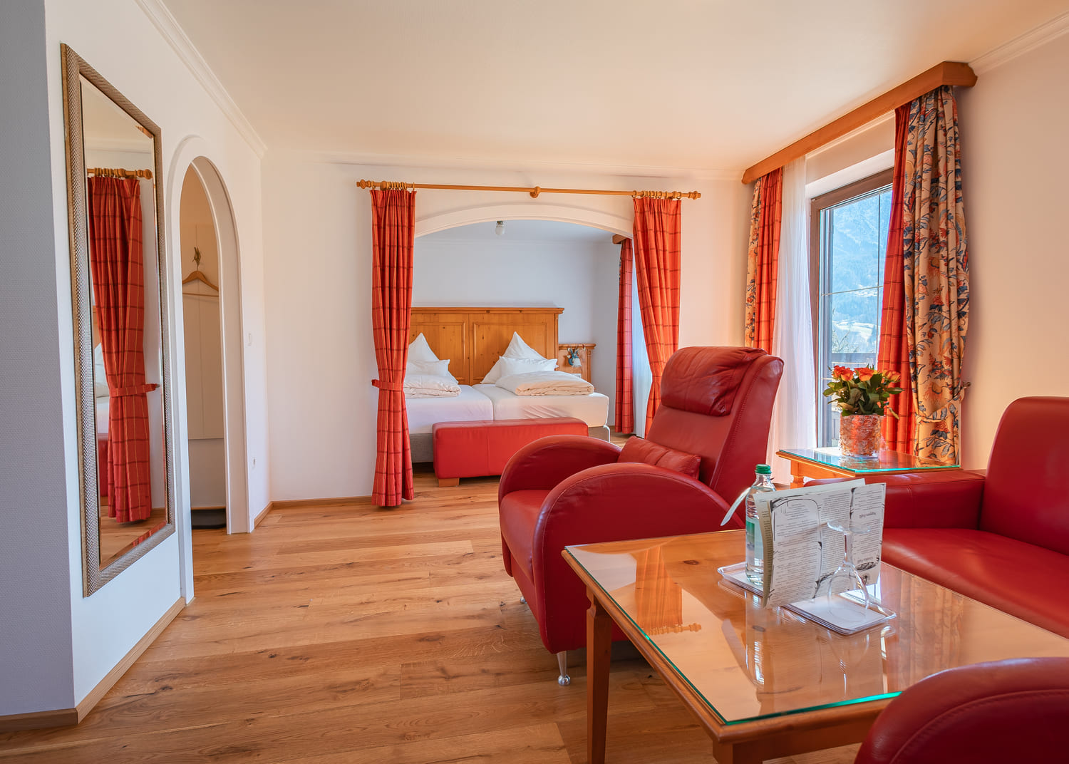 Helle Hotel-Suite mit roten Ledersesseln, Holzboden, Glas-Couchtisch und durch rote Vorhänge abgetrenntem Doppelbett sowie Fenster mit Bergblick.