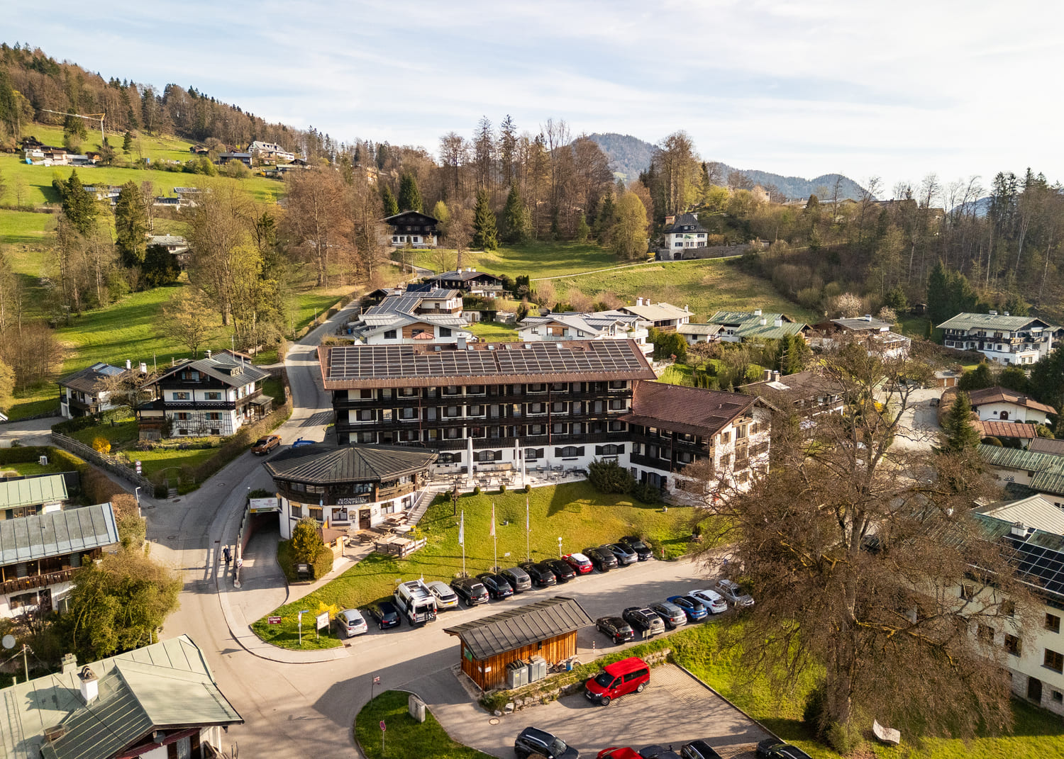 Luftaufnahme eines Hotels mit Solarpaneelen und Parkplätzen in einem kleinen Alpenort, umgeben von Häusern und grünen Hügeln.