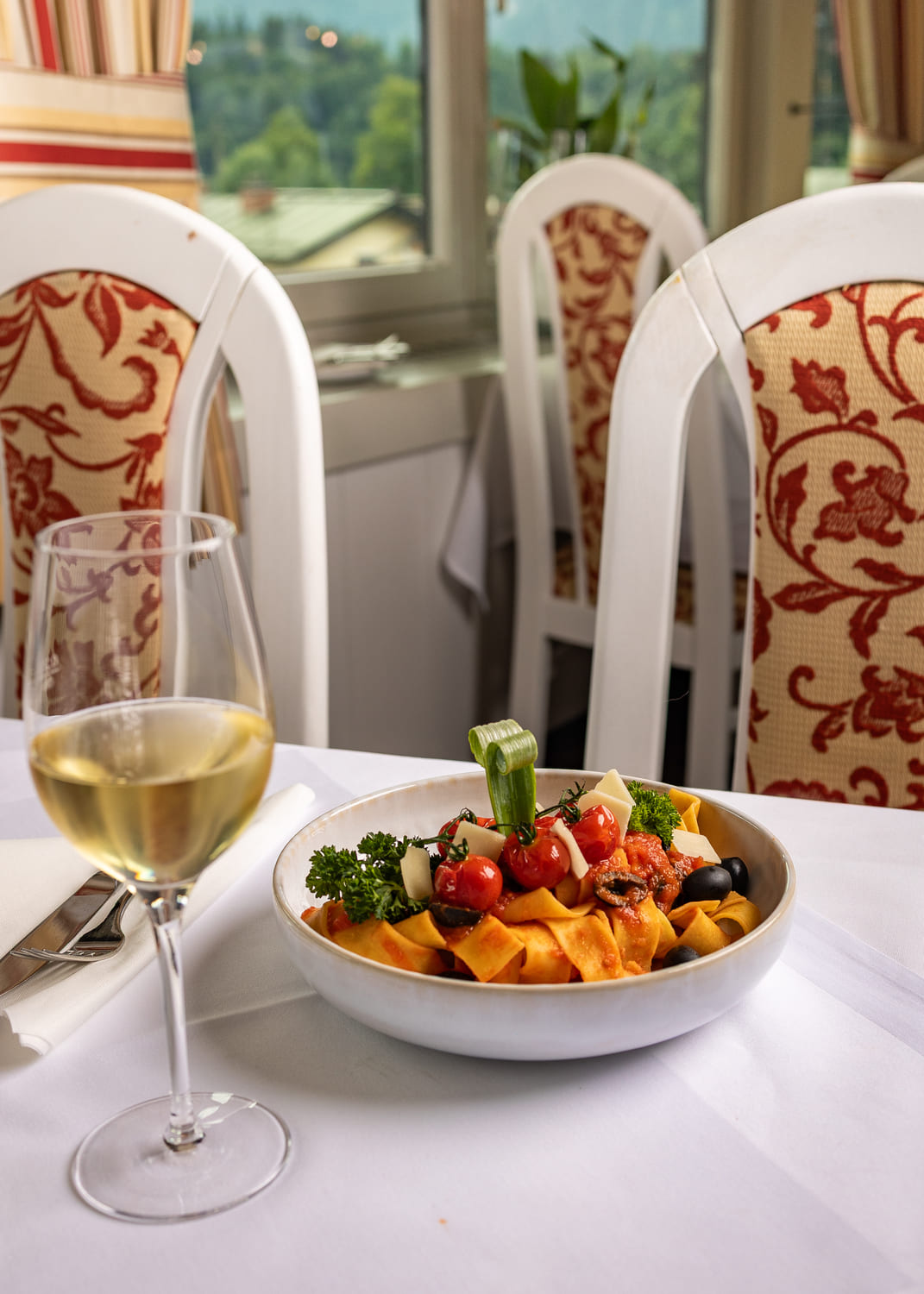 Teller Bandnudeln mit Tomatensauce, Kirschtomaten, Oliven, Petersilie und Käse neben einem Glas Weißwein in elegantem Restaurant.