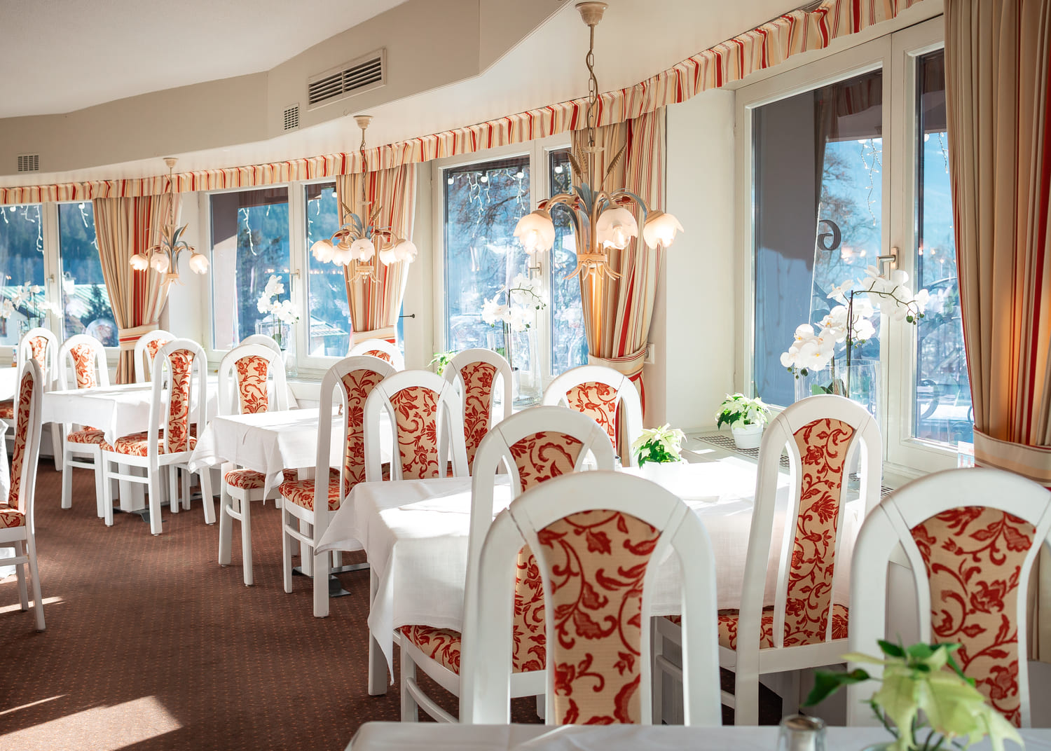 Heller Speisesaal mit weißen Stühlen in rotem Muster, weiß gedeckten Tischen, großen Fenstern und Hängelampen.