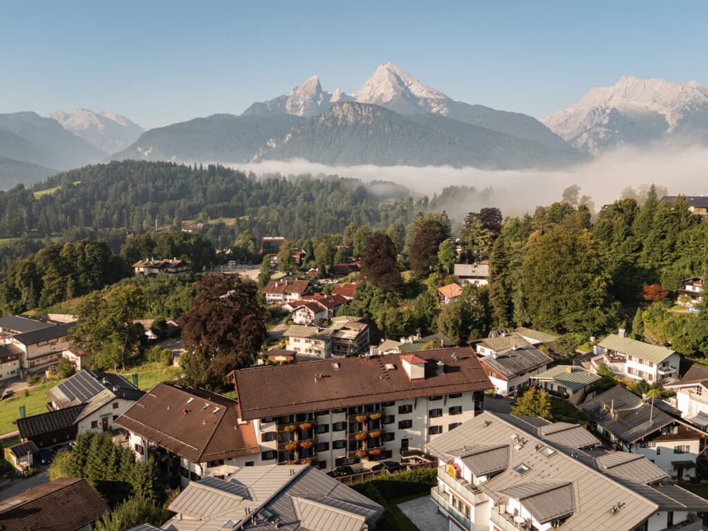 Alpendorf mit Häusern und Blumenkästen, Morgennebel über bewaldeten Hügeln und schneebedeckten Bergen im Hintergrund.