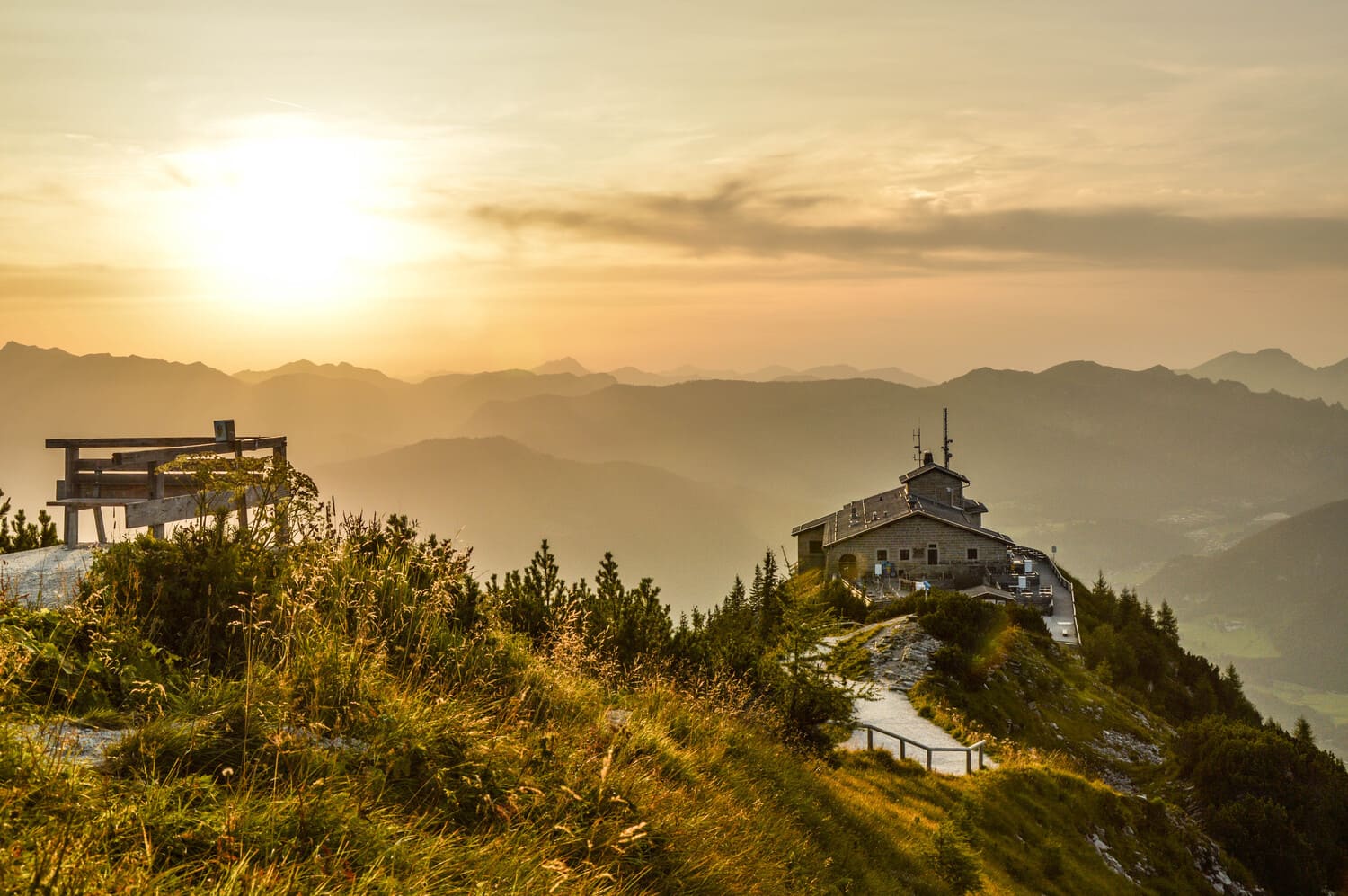 Berggasthaus auf felsigem Grat mit Weg und Holzbänken im Vordergrund, goldener Sonnenuntergang über nebeligen Bergketten.