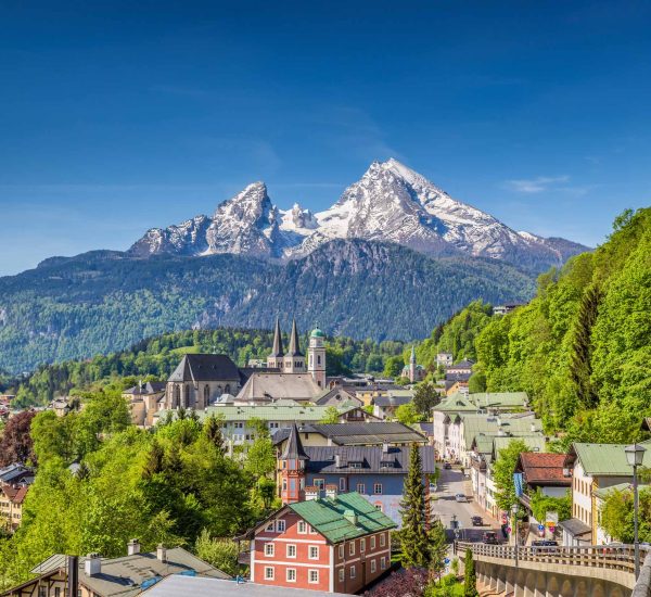 Kleinstadt mit Kirchtürmen und bunten Häusern am Hang, vor schneebedeckten Alpengipfeln und strahlend blauem Himmel.