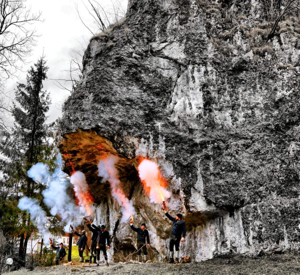 Menschen unter einem Felsüberhang halten brennende Fackeln; orange Flammen und dichter weißer Rauch steigen auf.