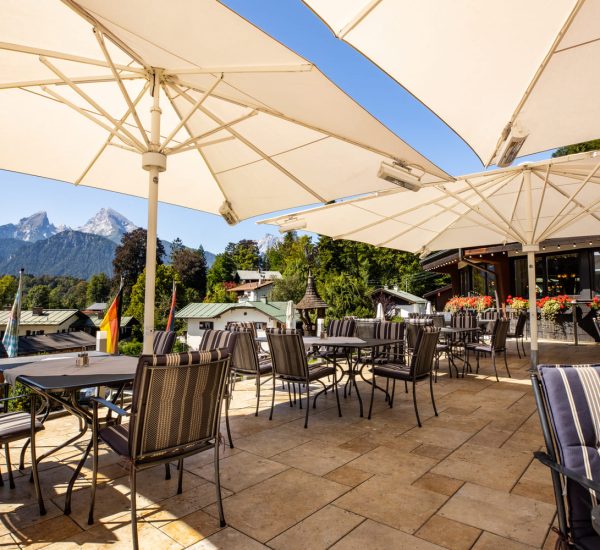 Sonnige Terrasse mit gestreiften Sitzpolstern und großen weißen Sonnenschirmen, Blick auf bewaldete Hügel und Berge.