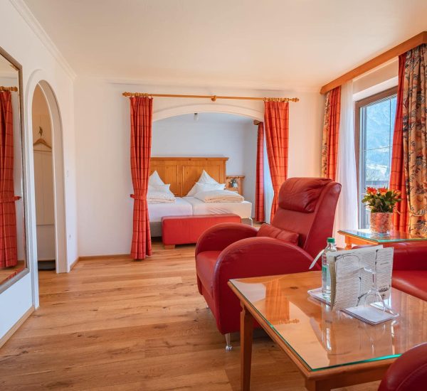 Helle Hotel-Suite mit roten Ledersesseln, Holzboden, Glas-Couchtisch und durch rote Vorhänge abgetrenntem Doppelbett sowie Fenster mit Bergblick.