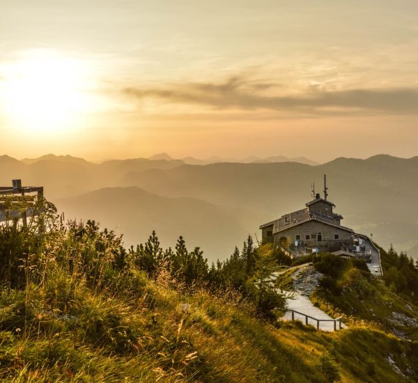 Berggasthaus auf felsigem Grat mit Weg und Holzbänken im Vordergrund, goldener Sonnenuntergang über nebeligen Bergketten.