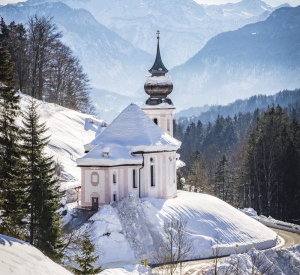 Barocke Kirche mit Zwiebelturm und schneebedecktem Dach an einer kurvigen Bergstraße, im Hintergrund verschneite Alpen.