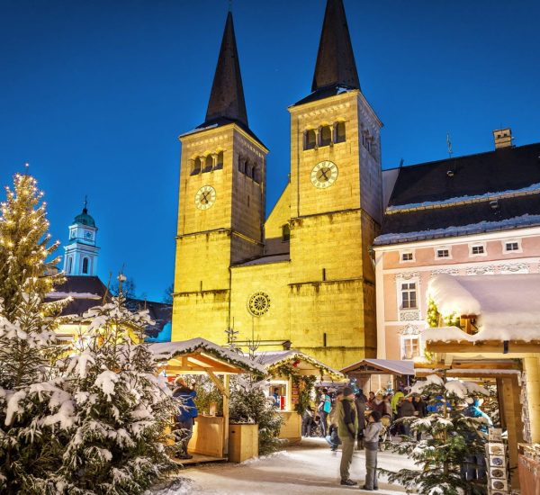 Weihnachtsmarkt bei Nacht vor einer beleuchteten Doppelkirche, verschneite Tannen, Lichter und Holzstände.