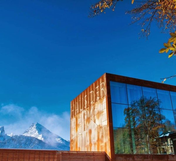 Rostfarbener Würfelbau mit großen Glasfenstern und geprägter Aufschrift, die Bäume spiegeln sich, vor schneebedeckten Bergen.