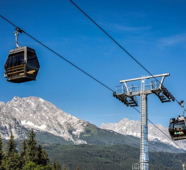 Zwei moderne Seilbahnkabinen an einer Stahlstütze über Nadelwald vor schneebedeckten Alpen und blauem Himmel.