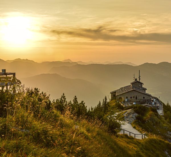 Berggasthaus auf einem Grat bei goldenem Sonnenuntergang, Holzbank im Vordergrund und nebelige Bergsilhouetten.