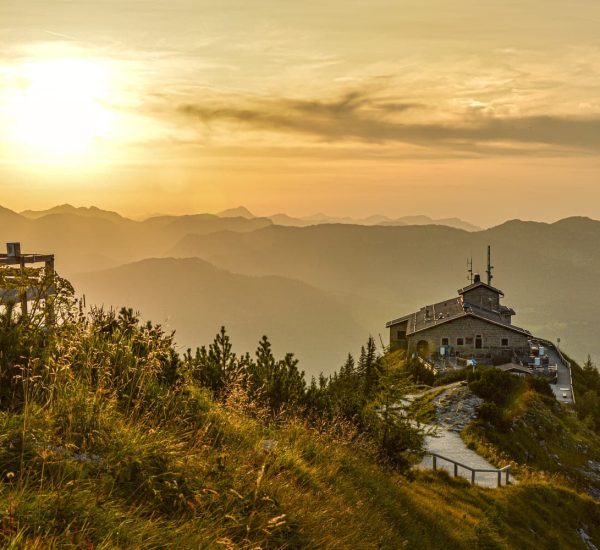 Berggasthaus auf einem Grat bei goldenem Sonnenuntergang, Holzbank im Vordergrund und nebelige Bergsilhouetten.