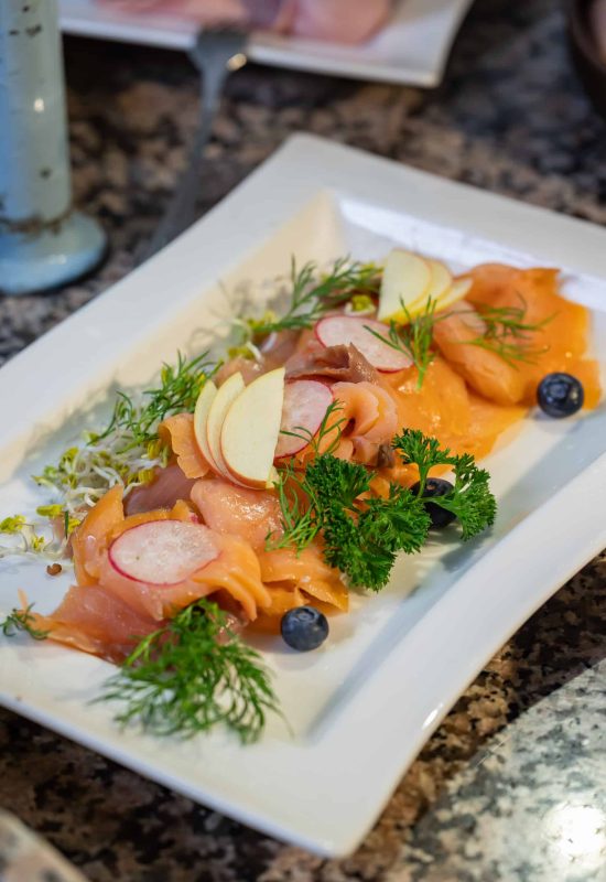 Auf einem weißen Teller angerichtene Räucherlachsscheiben mit Apfelscheiben, Radieschen, Dill, Petersilie und Blaubeeren.