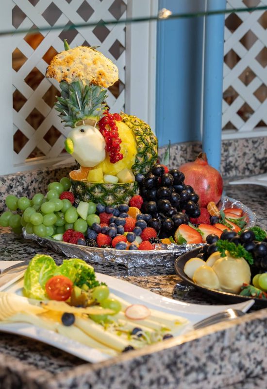 Buffetplatte mit kunstvoll geschnitzter Ananasfigur, umgeben von Trauben, Beeren, Erdbeeren, Granatapfel und Käseplatten auf Granittheke.