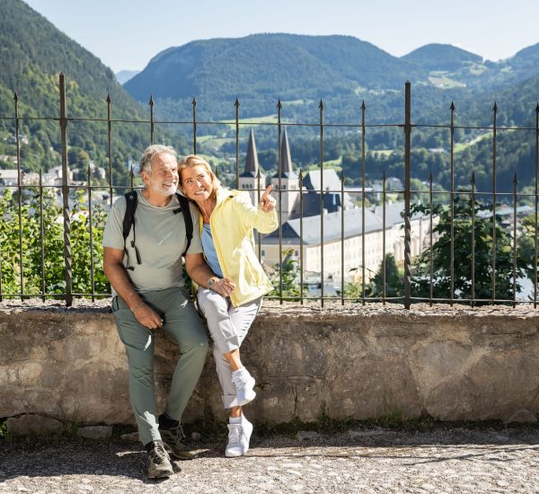 Älteres Paar mit Rucksäcken sitzt auf einer Steinmauer; die Frau zeigt lächelnd auf ein Bergtal mit Kirchturm im Hintergrund.