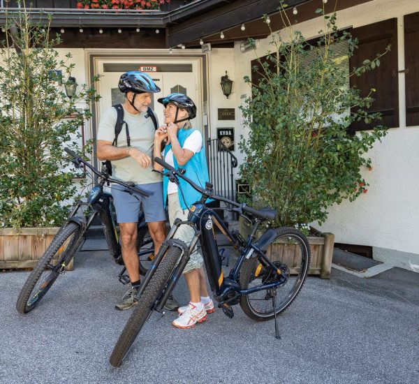 Älteres Paar mit Helmen befestigt die Kinnriemen an ihren E‑Bikes vor einem Hoteleingang.