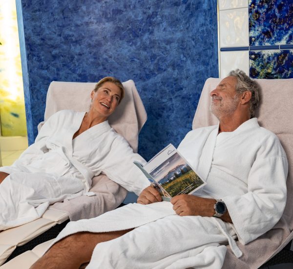 Älteres Paar in weißen Bademänteln entspannt auf Liegen im Wellnessbereich, Mann liest Broschüre vor blauer Wandfliese.