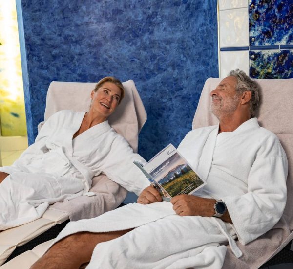 Älteres Paar in weißen Bademänteln entspannt auf Liegen im Wellnessbereich, Mann liest Broschüre vor blauer Wandfliese.