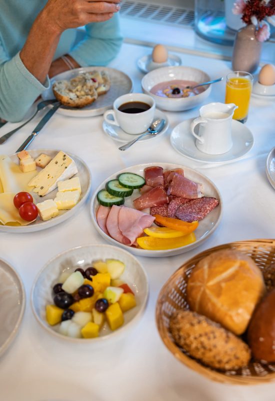 Frühstückstisch mit Wurst- und Käseplatten, Gurken- und Paprikascheiben, Obstsalat, Brötchen, Kaffee und Orangensaft.