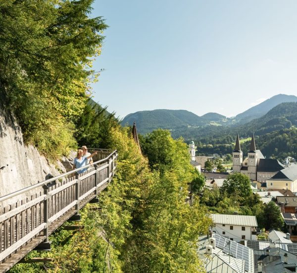 Paar auf hölzerner Steiganlage am Felsrand blickt über grüne Täler auf eine Alpenstadt mit Doppelturmkirche und Bergen.