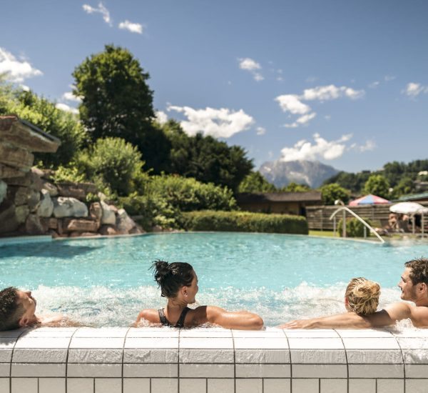 Vier Personen lehnen entspannt am Beckenrand eines Außenpools, im Hintergrund Grün, Felsen und Berge unter blauem Himmel.