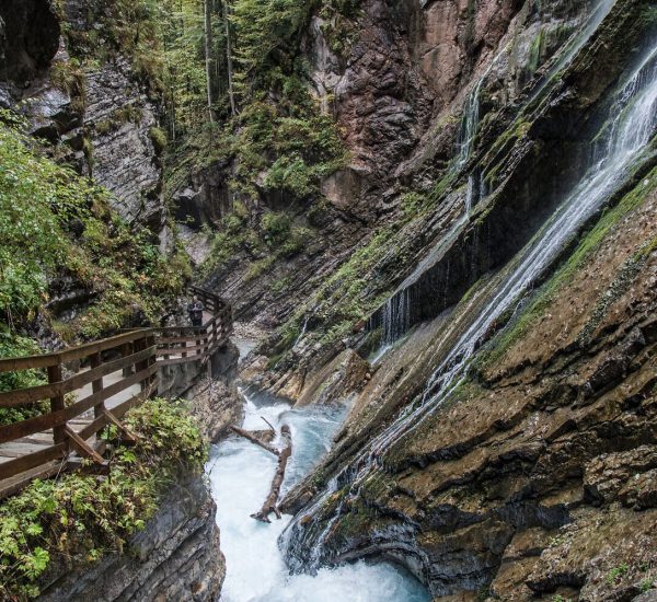 Holzsteg an einer engen Felsenschlucht mit Wasserfall über moosige Gesteinsstufen und reißendem, schäumendem Fluss.