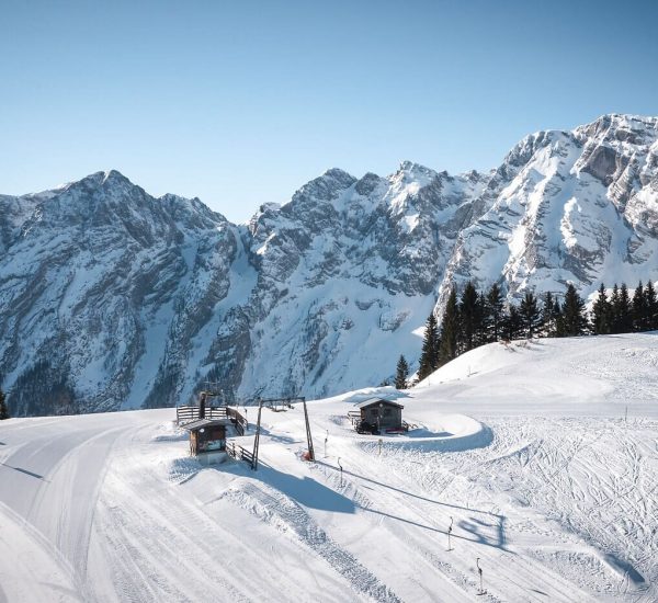 Verschneite Berglandschaft mit Skipiste, zwei Hütten, Schlepplift und Tannen vor schroffen, schneebedeckten Gipfeln.