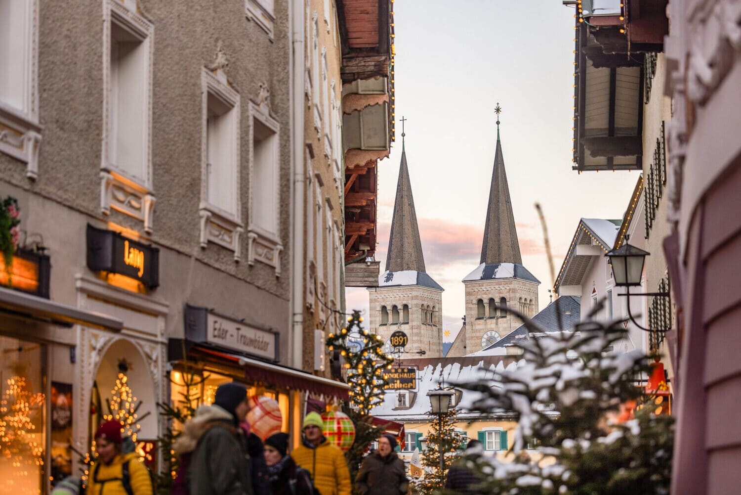 Belebte Weihnachtsmarkt-Gasse mit festlich geschmückten Ständen, Besuchern und zwei hohen Kirchtürmen im Hintergrund.