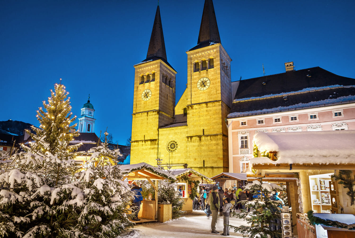 Weihnachtsmarkt bei Nacht vor einer beleuchteten Doppelkirche, verschneite Tannen, Lichter und Holzstände.