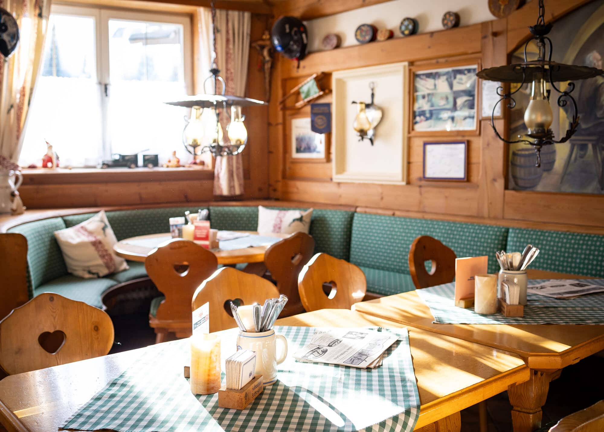 Gemütliche Wirtsstube mit holzernen Tischen und Herz-ausgeschnittenen Stühlen, karierten Tischläufern und grüner Eckbank.