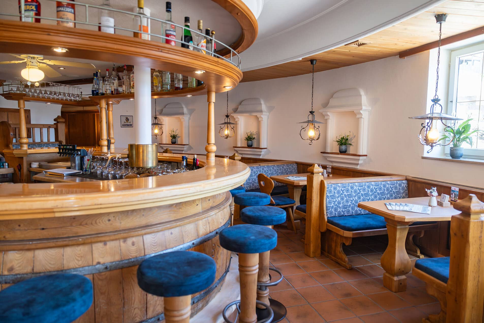 Gemütliche Holzbar mit runden Barhockern in blau, eingelassenen Sitznischen, hängenden Laternen und Flaschenregal über dem Tresen.