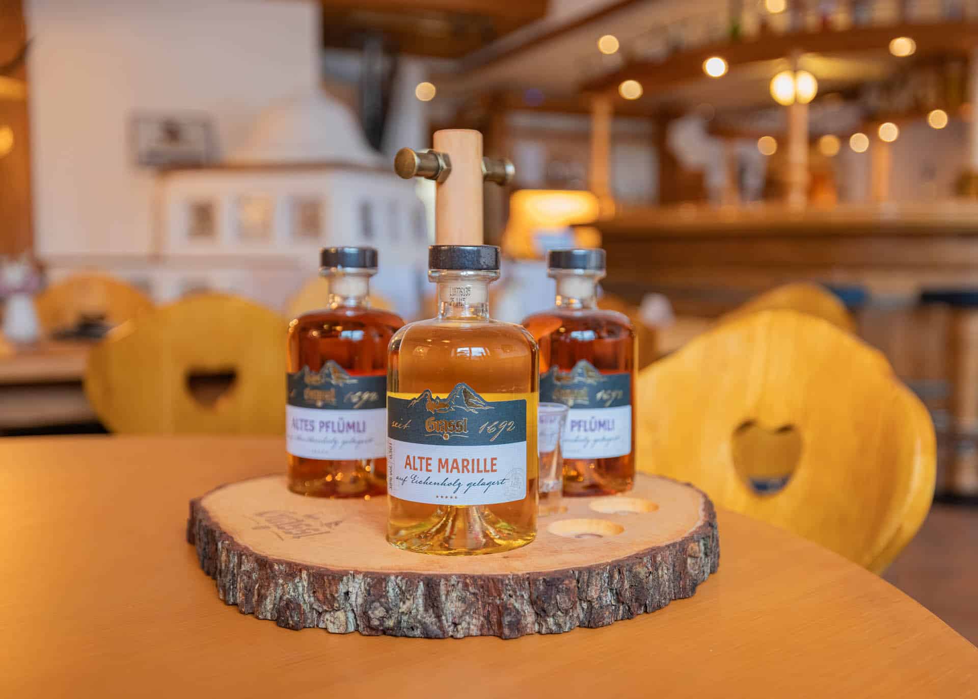 Drei Flaschen Obstbrand auf einer Baumscheibe im Gasthaus, im Vordergrund Etikett "Alte Marille", Holzstühle mit Herz-Ausschnitt im Hintergrund
