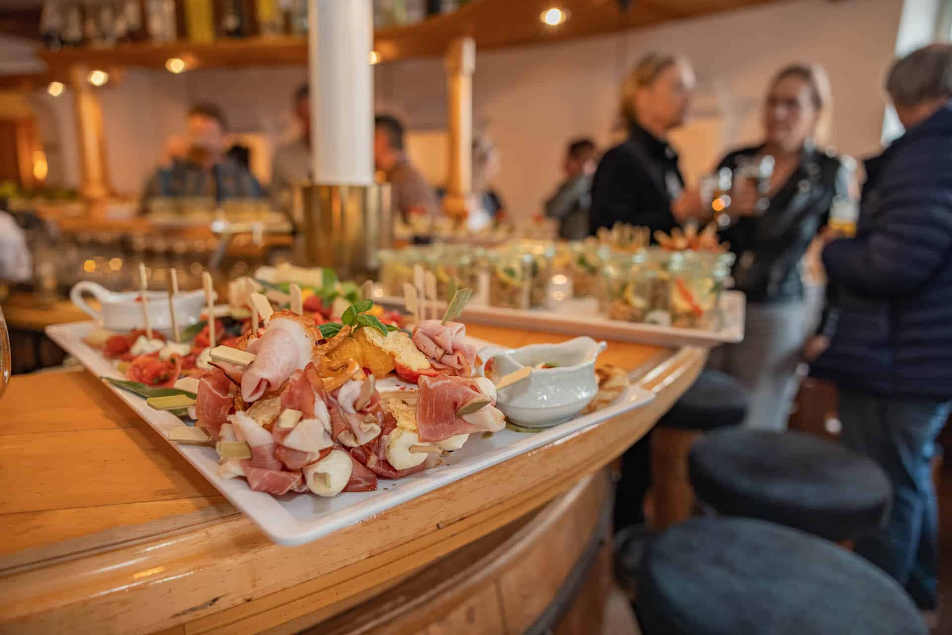 Platte mit aufgespießtem Prosciutto, Mozzarella und Obst auf Bartresen, unscharfe Gäste im Hintergrund bei einem Empfang.