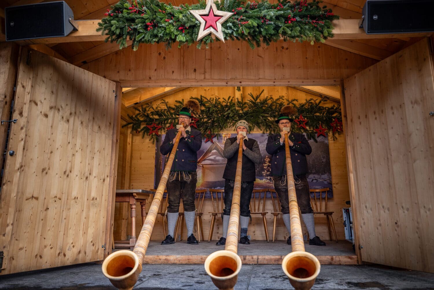 Drei Musiker in Tracht blasen lange Alphörner aus einer hölzernen Bude, geschmückt mit Tannengirlanden und einem roten Stern.