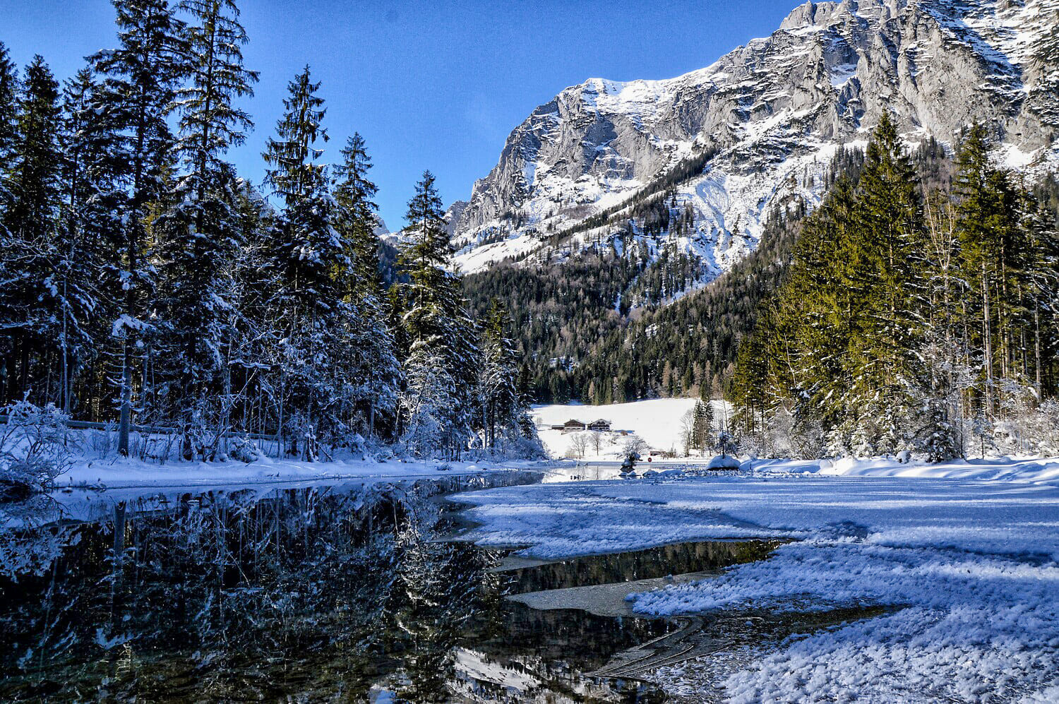 Verschneite Tannen am zugefrorenen See mit Spiegelung, schneebedeckte Berge und kleine Hütten im Hintergrund.