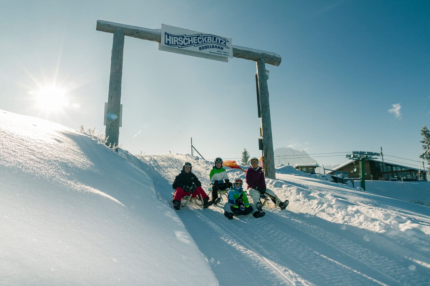 Vier Personen auf Holzschlitten unter einem Holzbogen mit Schild Hirscheckblitz Rodelbahn auf sonniger, verschneiter Piste.
