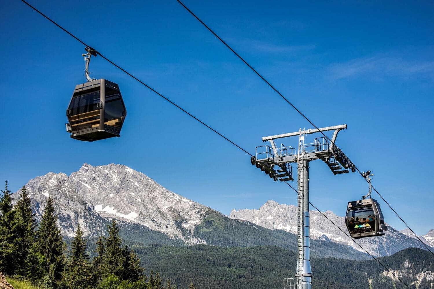 Zwei moderne Seilbahnkabinen an einer Stahlstütze über Nadelwald vor schneebedeckten Alpen und blauem Himmel.