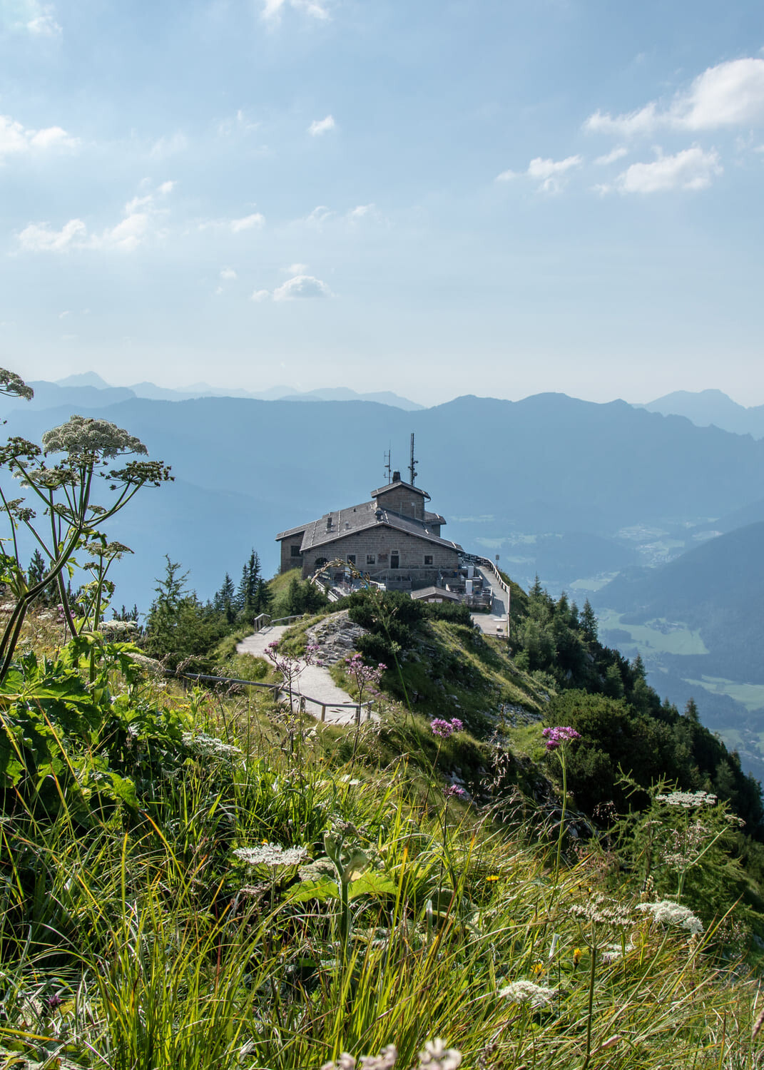 Berghütte auf einem grasigen Gipfelrücken mit Wanderweg, Wildblumen im Vordergrund und blauen Bergketten im Dunst.