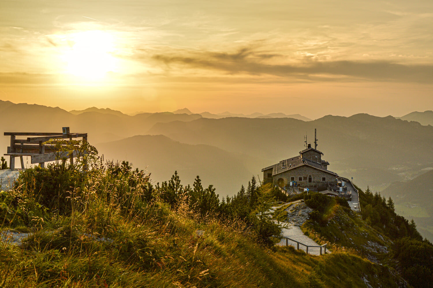 Berggasthaus auf einem Grat bei goldenem Sonnenuntergang, Holzbank im Vordergrund und nebelige Bergsilhouetten.