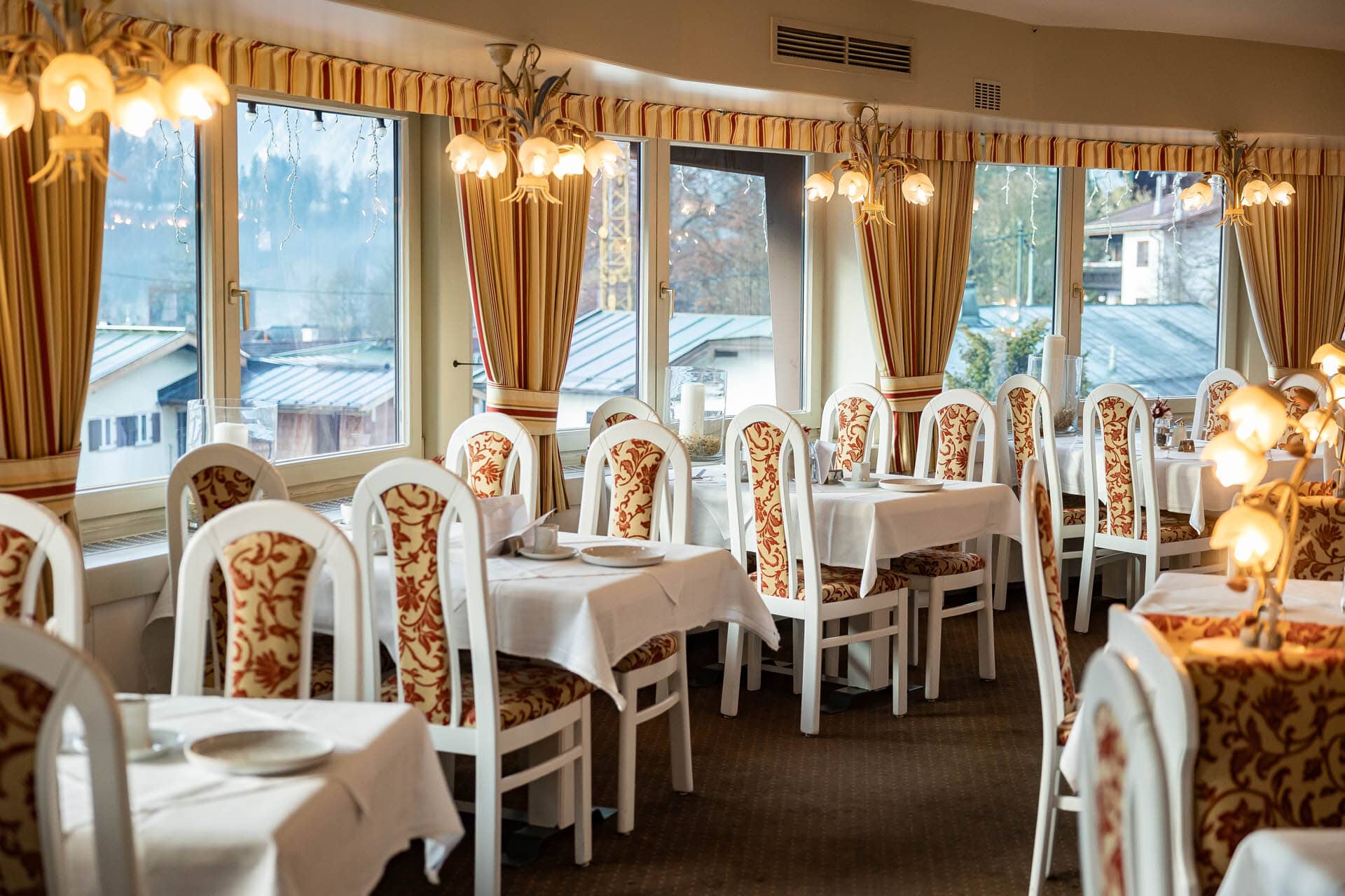 Helles, elegant gedecktes Restaurant mit weißen Tischen, gestreiften Vorhängen und gemusterten Polsterstühlen an großen Fenstern.