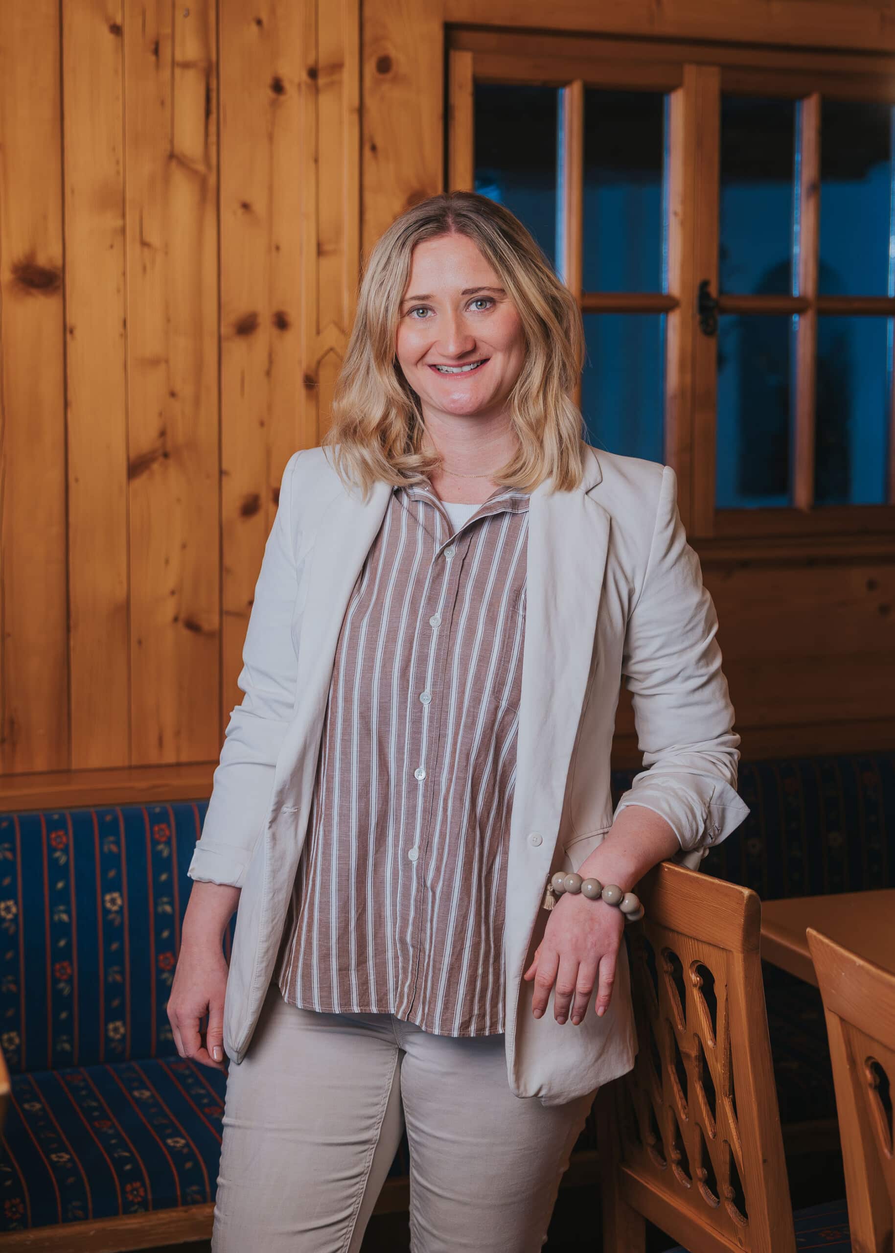 Blonde Frau in hellbeiger Jacke und gestreiftem Hemd, lächelnd, lehnt an einem Holzstuhl in rustikalem Raum.