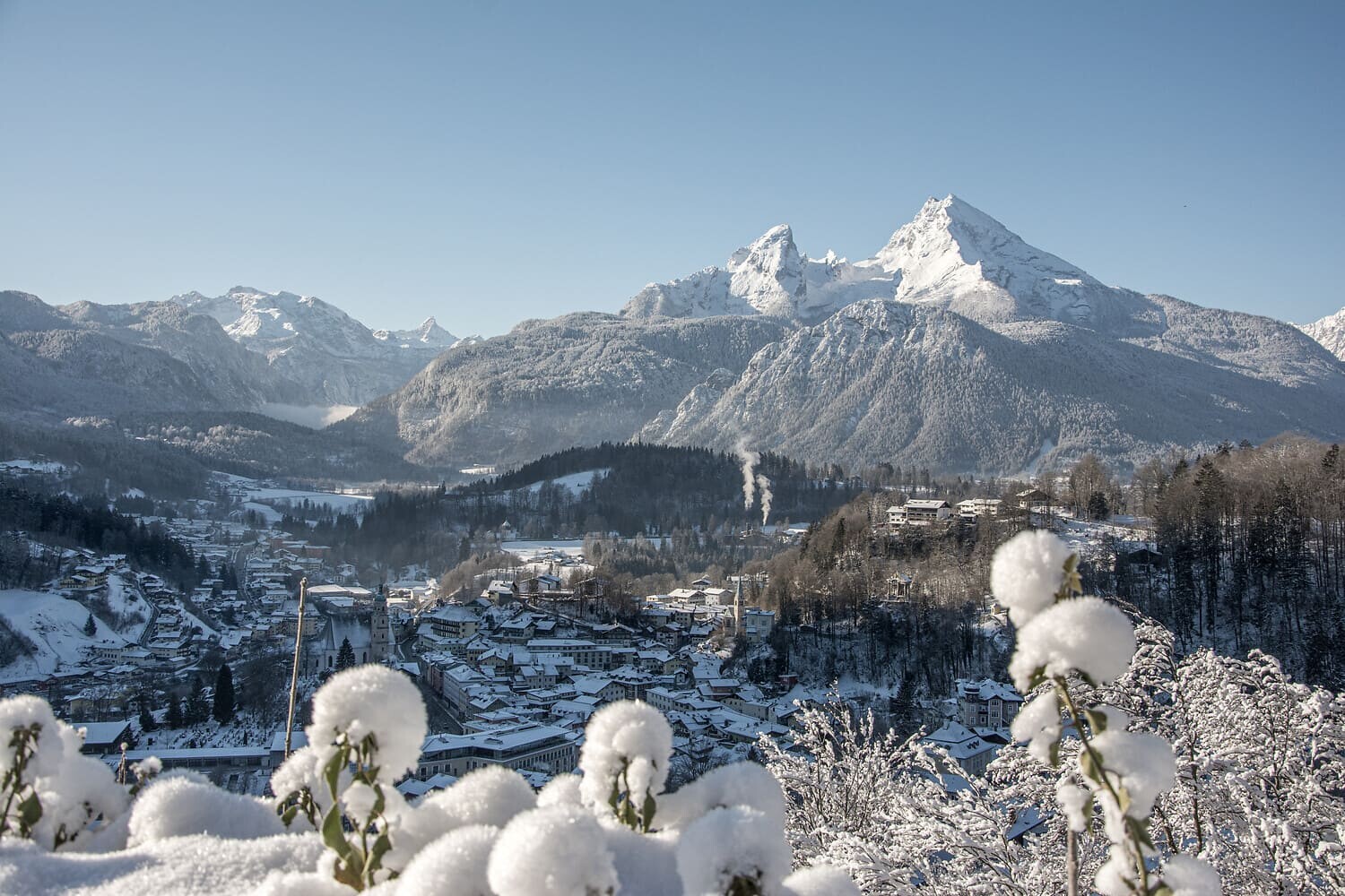 Verschneites Dorf im Tal vor schneebedeckten Berggipfeln bei klarem Winterhimmel, im Vordergrund verschneite Pflanzen.