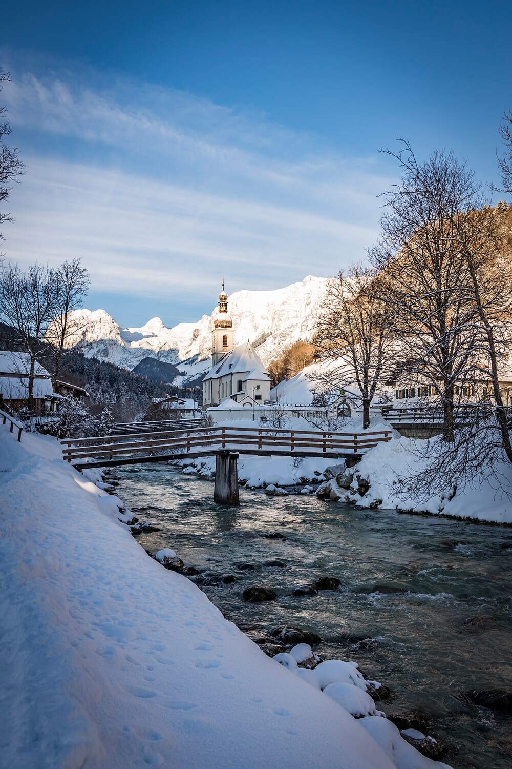 Verschneites Bergdorf mit Holzbrücke über Bach und Kirche mit Zwiebelturm vor schneebedeckten Alpen.