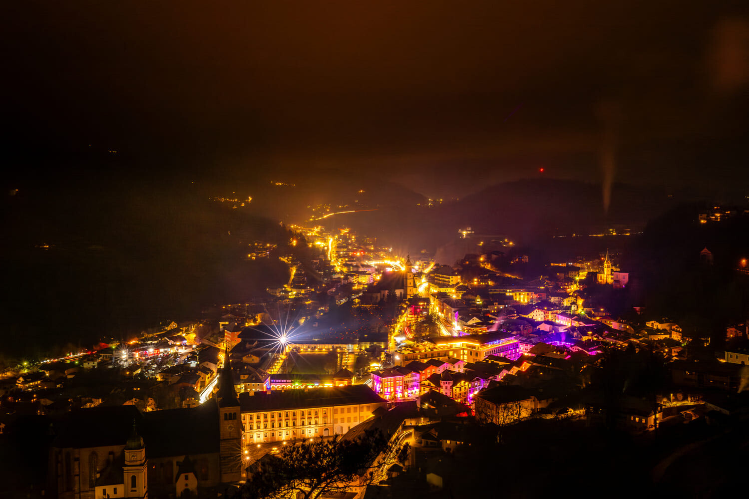 Nächtliche Kleinstadt im Tal mit warmen Straßenlichtern, bunten Gebäudebeleuchtungen, Kirchturm und nebelverhangenen Hügeln.