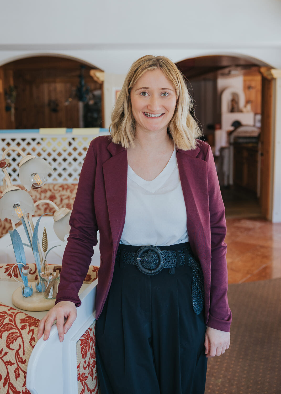 Lächelnde blonde Frau in burgunderfarbener Jacke und weißem Top, steht drinnen an gemustertem Sofa neben Lampe.