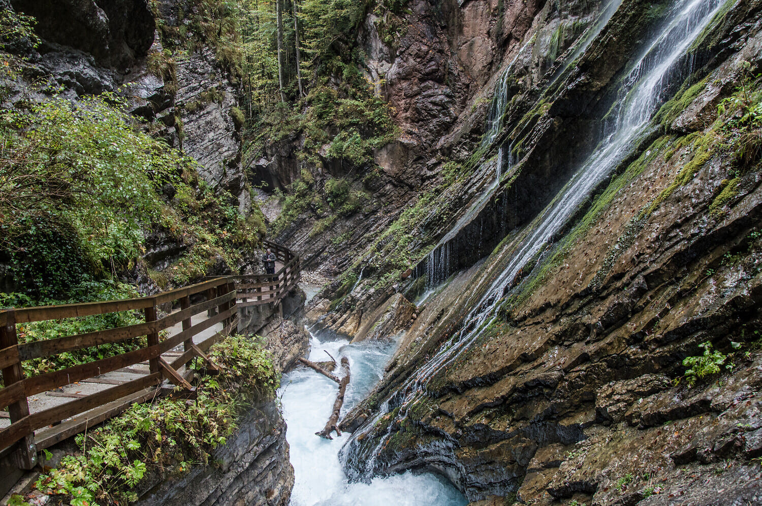 Holzsteg an einer engen Felsenschlucht mit Wasserfall über moosige Gesteinsstufen und reißendem, schäumendem Fluss.